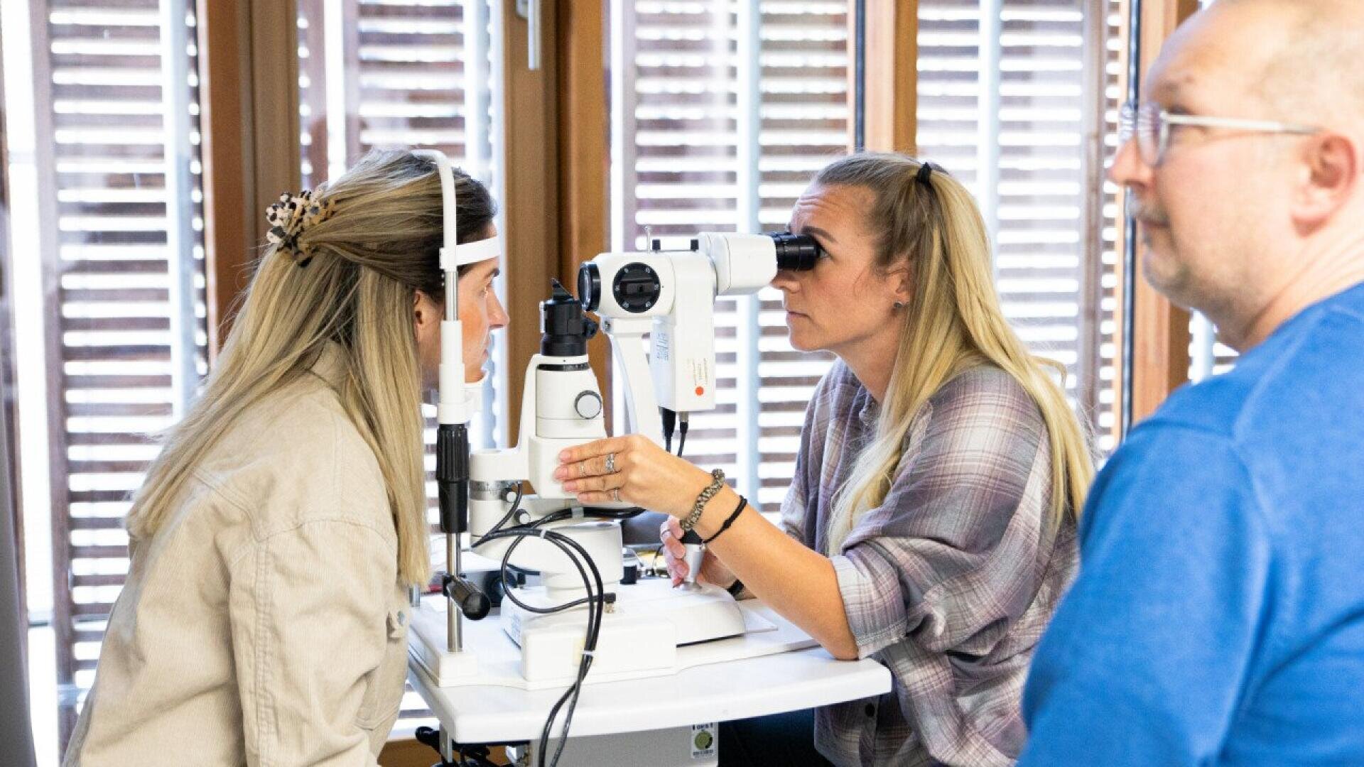 A woman examines another womans eyes using a slit lamp machine in a bright room, while a man in glasses observes nearby.