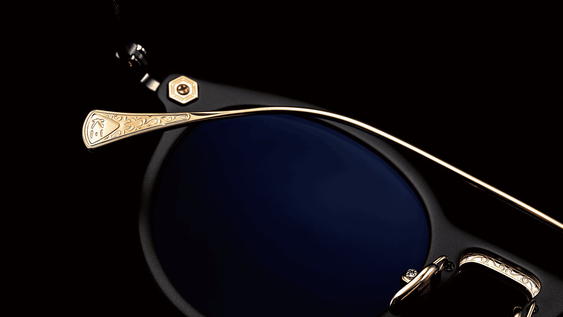 Nahaufnahme einer Sonnenbrille mit dunkelblauen Gläsern und einem goldenen, kunstvoll gravierten Bügel vor schwarzem Hintergrund.