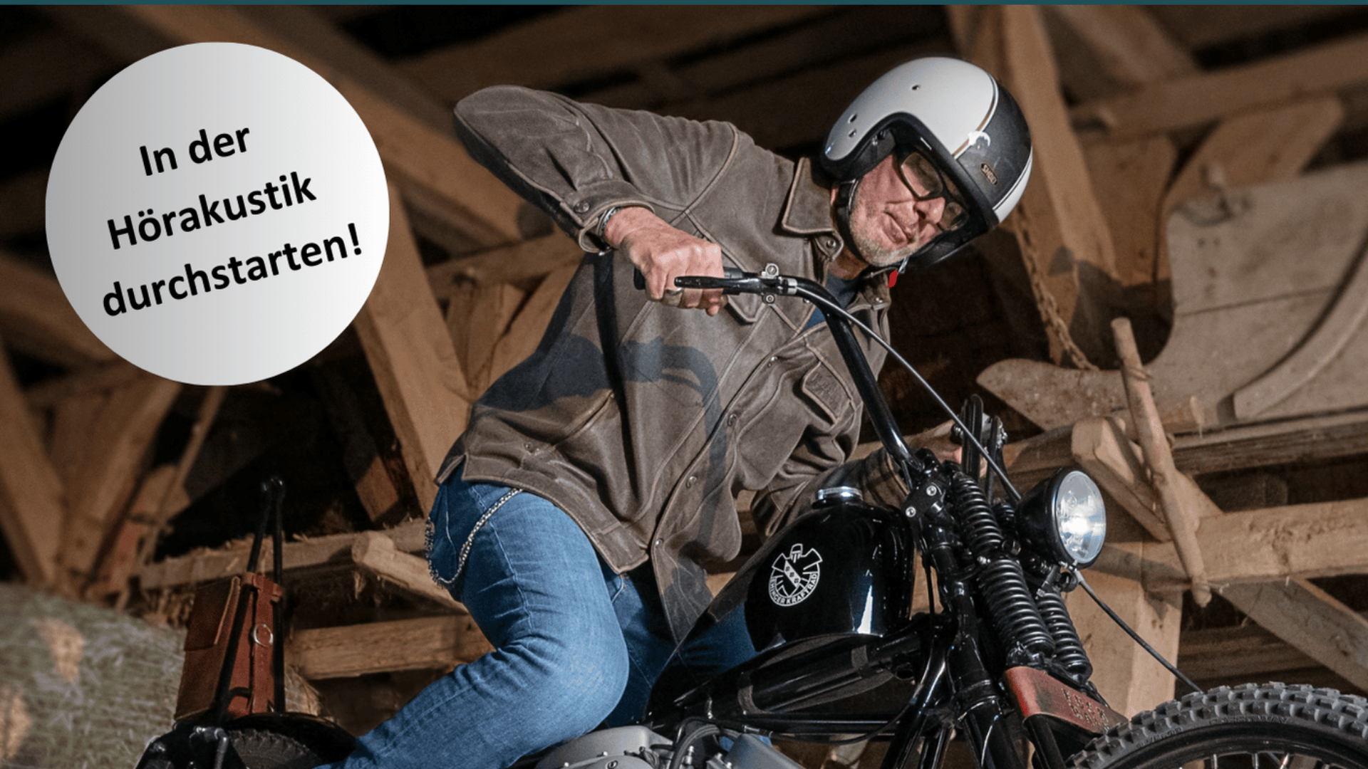 Ein Mann mit Helm und Brille sitzt auf einem Motorrad in einem rustikalen Holzgebäude. Auf einer kreisförmigen Grafik steht: „In der Hörakustik durchstarten!“.