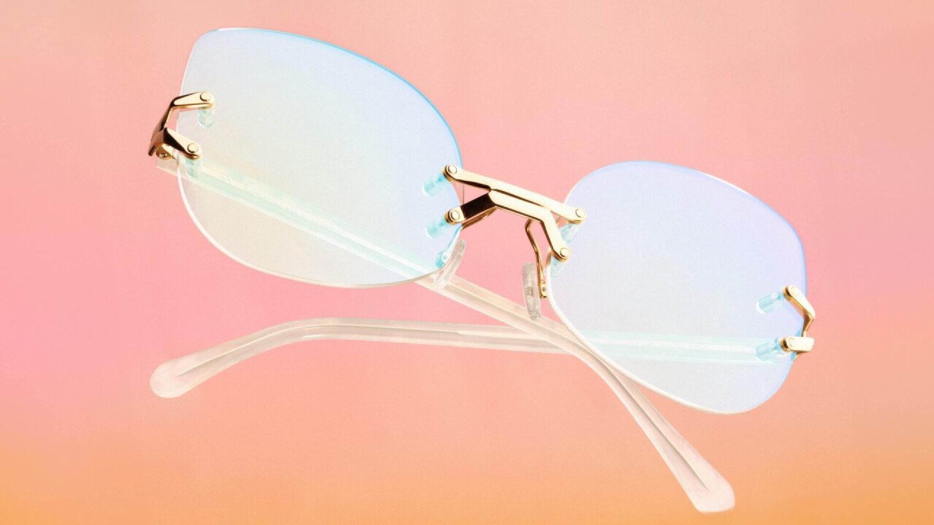 A pair of rimless, pastel-tinted glasses with gold hinges is shown against a pink and orange gradient background. The clear plastic arms are crossed below the lenses.