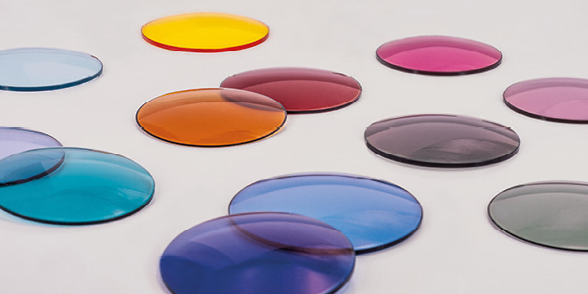 Round, flat lenses in various colors, including blue, orange, yellow, purple and red, are arranged on a bright surface. The lenses are translucent and evenly spaced.
