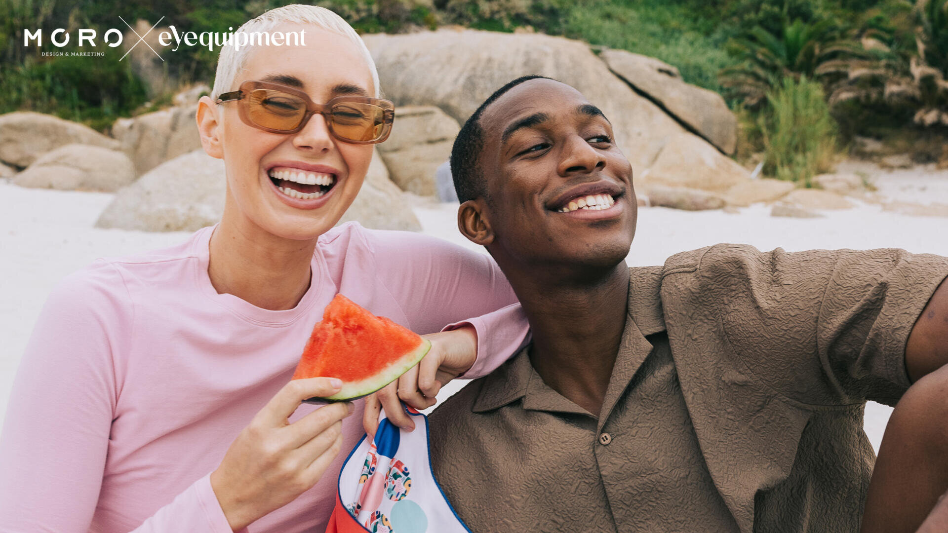 Eine lächelnde Frau mit kurzen blonden Haaren und Sonnenbrille hält ein Stück Wassermelone in der Hand, während sie neben einem lächelnden Mann an einem Sandstrand mit Felsen und Grün im Hintergrund sitzt.