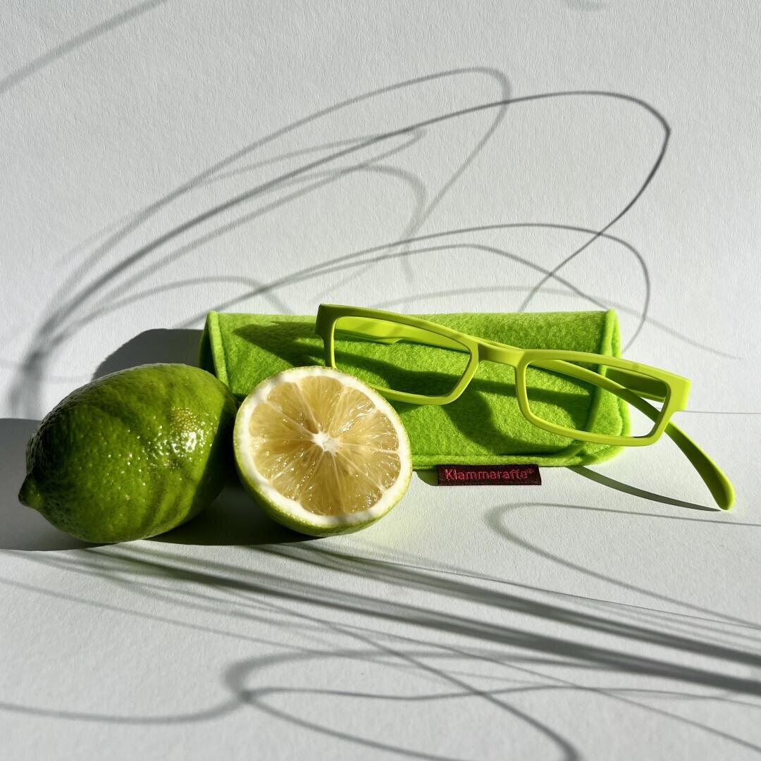 Eine hellgrüne Brille, ein dazu passendes grünes Brillenetui, eine ganze Limette und eine Limettenhälfte sind auf einer weißen Oberfläche angeordnet, wobei dramatische Schatten über die Szene geworfen werden.