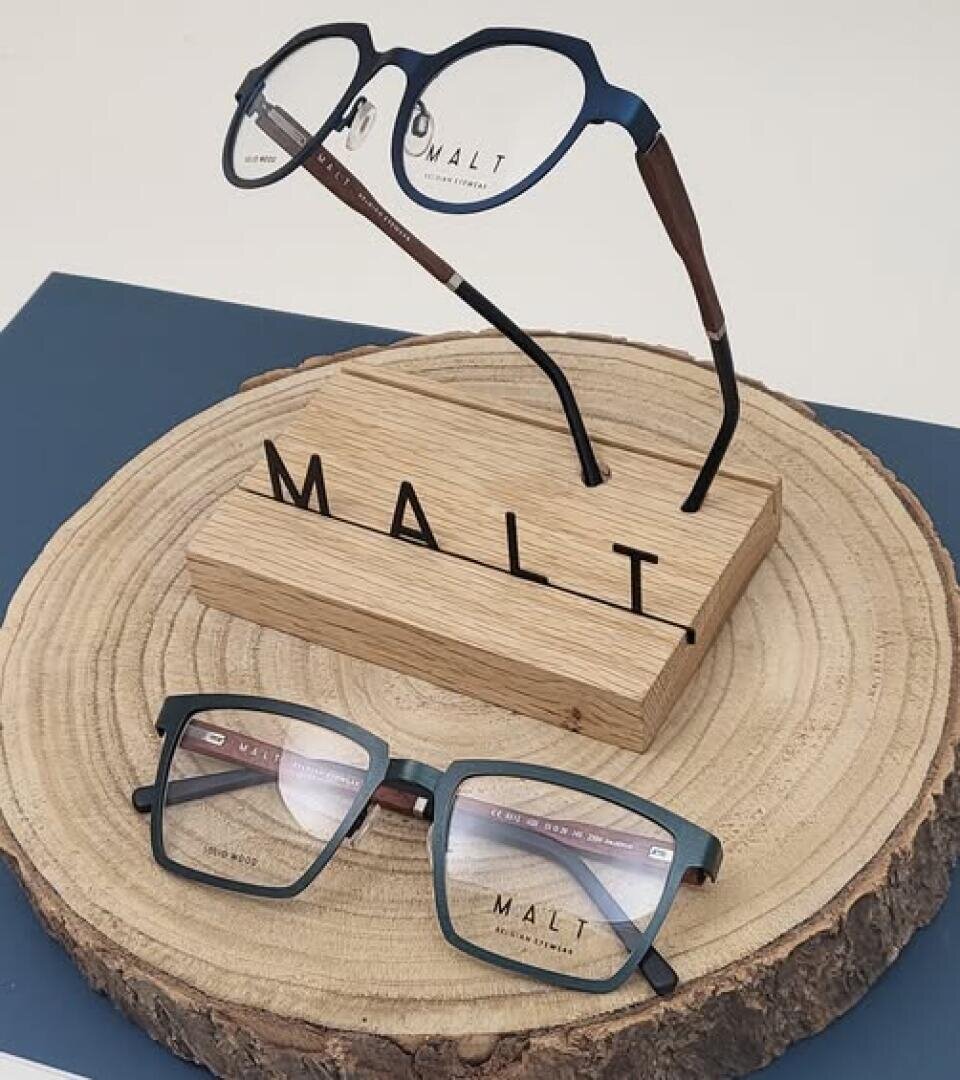 Auf einem runden Holzpodest sind zwei Brillen mit blauem Rand ausgestellt. Eine Brille steht aufrecht auf einem Holzständer mit der Aufschrift MALT, während die andere Brille flach davor liegt.