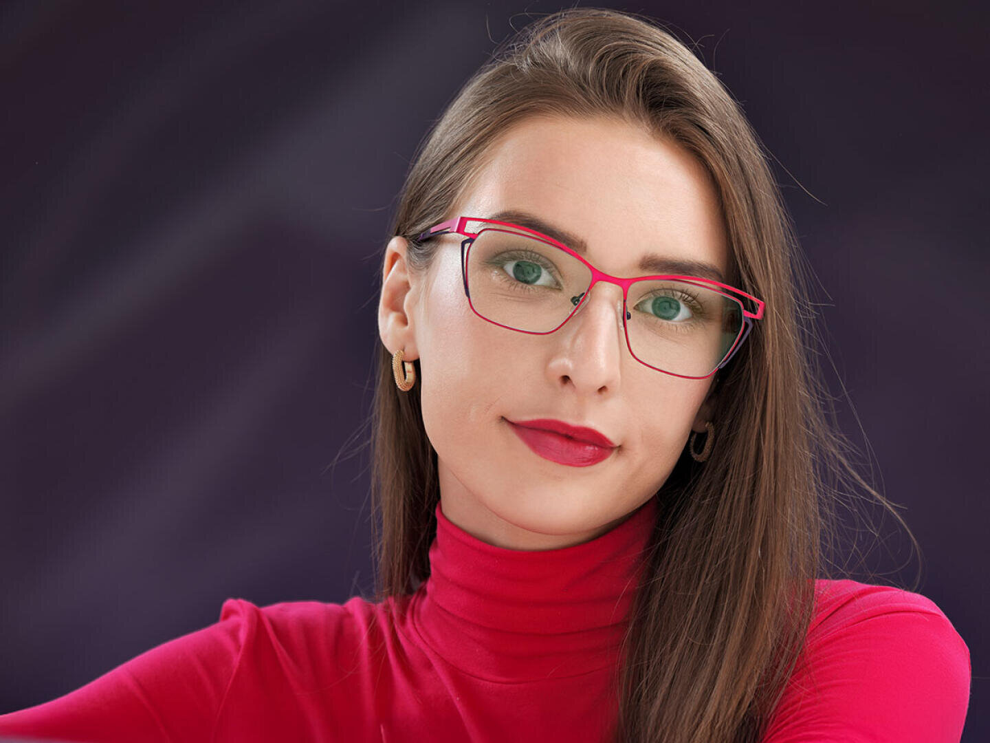 Eine Frau mit langen braunen Haaren, einer rotgerahmten Brille, einem roten Rollkragenpullover und goldenen Ohrringen posiert vor einem dunkelvioletten Hintergrund. Sie hat einen neutralen Gesichtsausdruck.