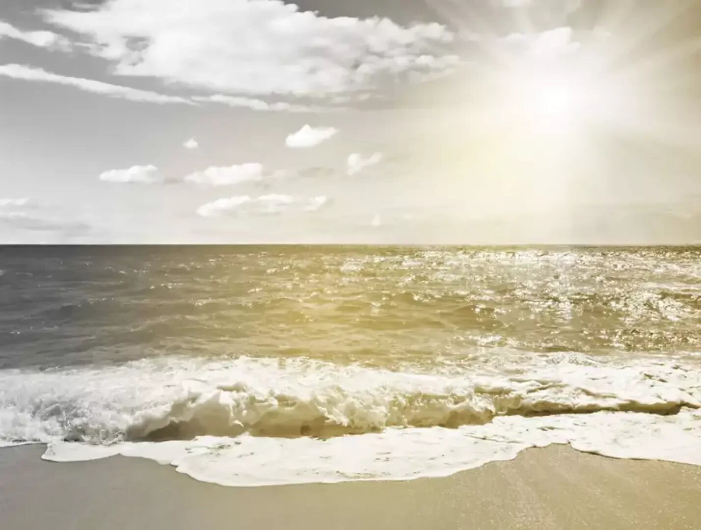 The sun shines brightly over a calm sea with gentle waves rolling onto a sandy beach. A few fluffy clouds are scattered in the sky, and the sunlight creates a golden reflection on the water.
