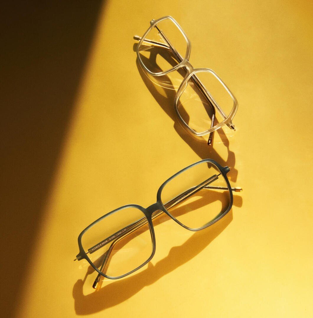 Two pairs of rectangular eyeglasses, one transparent and one dark-framed, are placed on a yellow surface with a diagonal shadow crossing the left side of the image.
