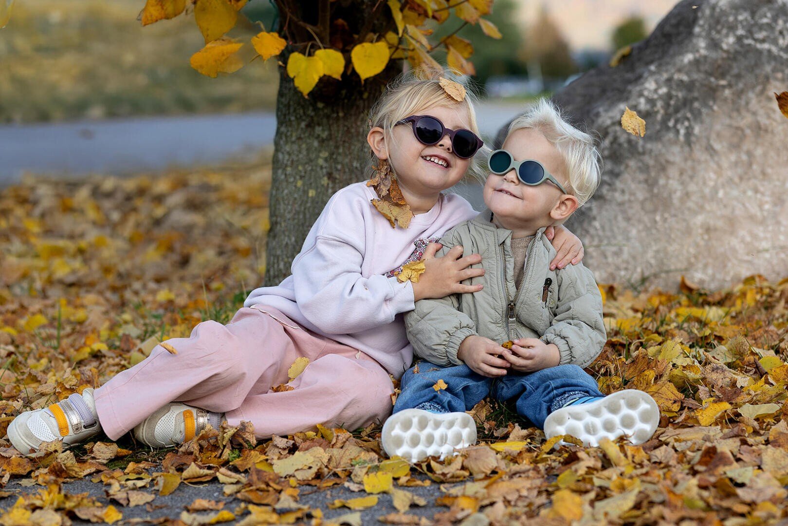 Two small children wearing sunglasses are sitting on the ground covered with autumn leaves, smiling and hugging each other near a tree. One is wearing pink trousers and a light-colored sweater, the other a beige jacket and blue trousers.