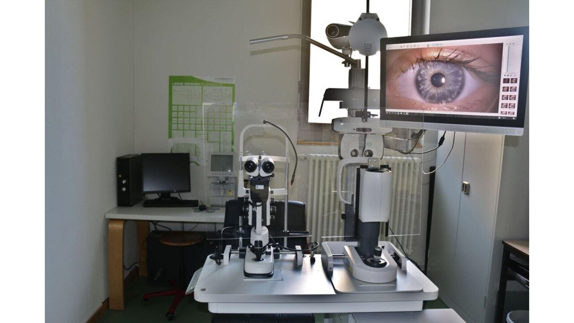 An eye examination room with ophthalmology equipment, including a slit lamp and a screen displaying a detailed image of a human eye. A desk with a computer is in the background.