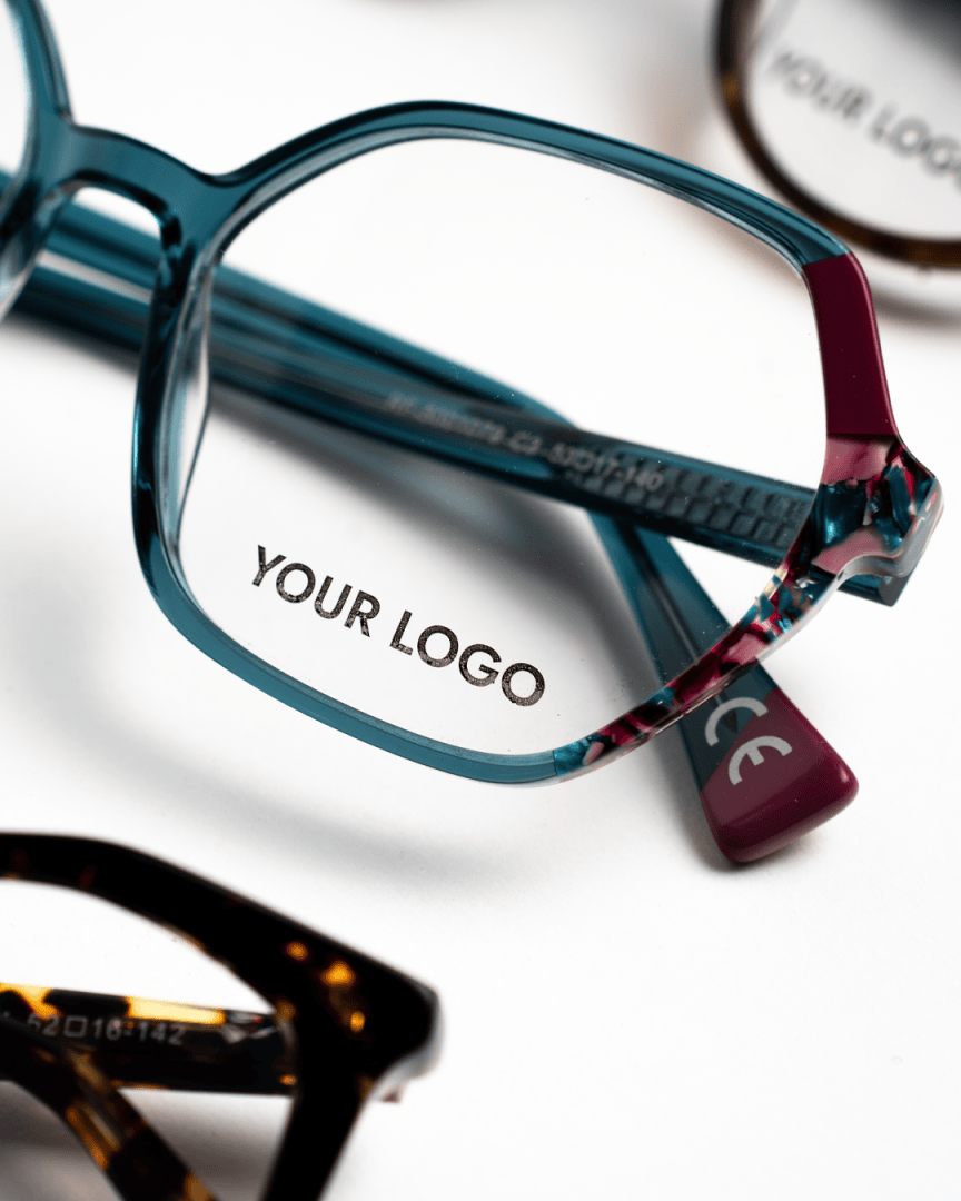 A close-up of eyeglass frames, with the words YOUR LOGO printed on the right lens. Other pairs of glasses are partially visible in the background.