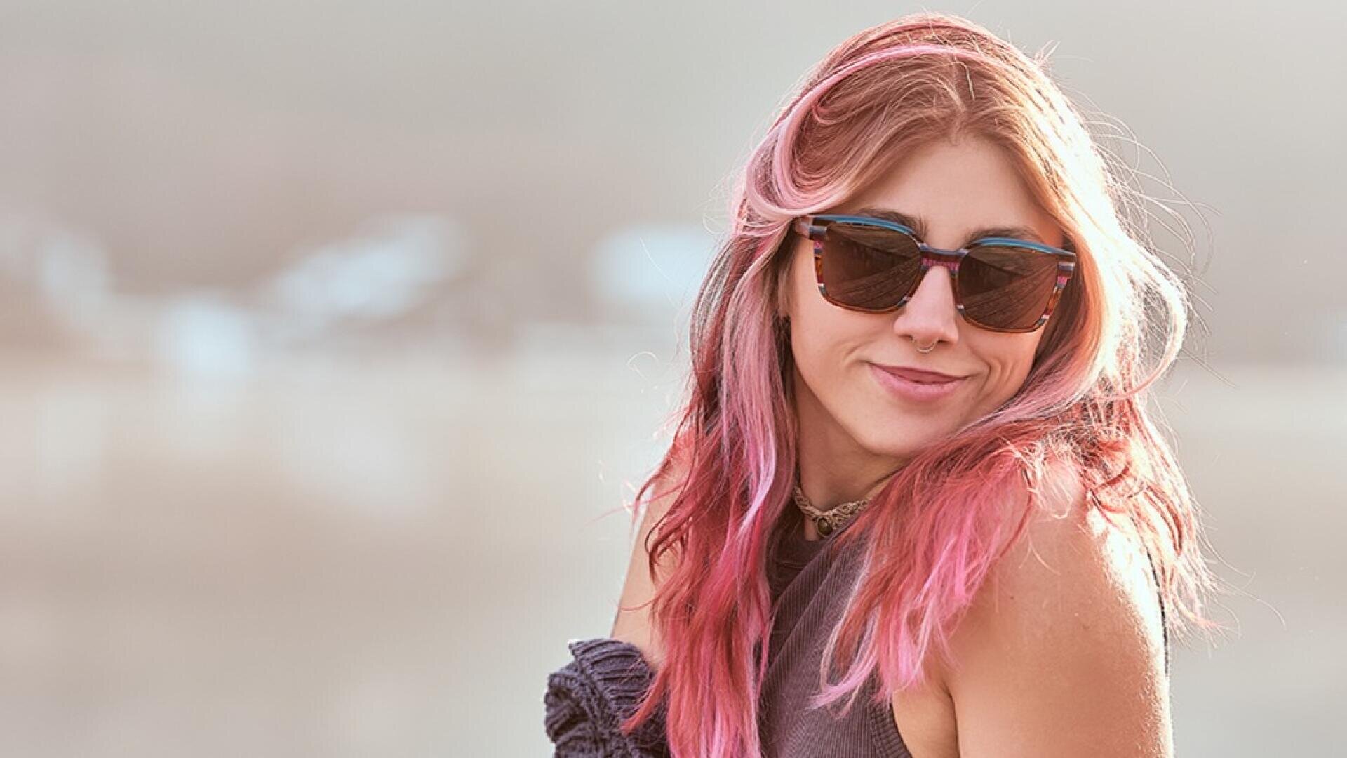 Eine junge Frau mit langen rosafarbenen Haaren, Sonnenbrille und ärmellosem Oberteil lächelt leicht, während sie im Freien bei sanftem natürlichem Licht über ihre Schulter schaut.