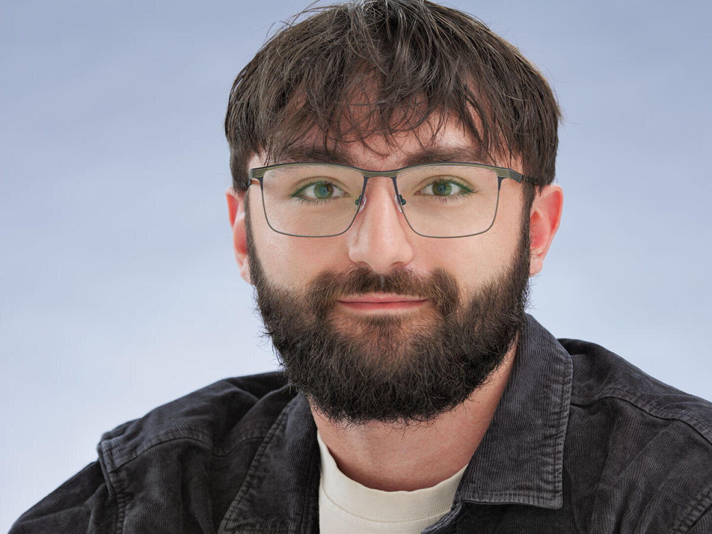 A young man with brown hair, a full beard and glasses, wearing a dark jacket over a light-colored shirt, looks into the camera against a light blue background.