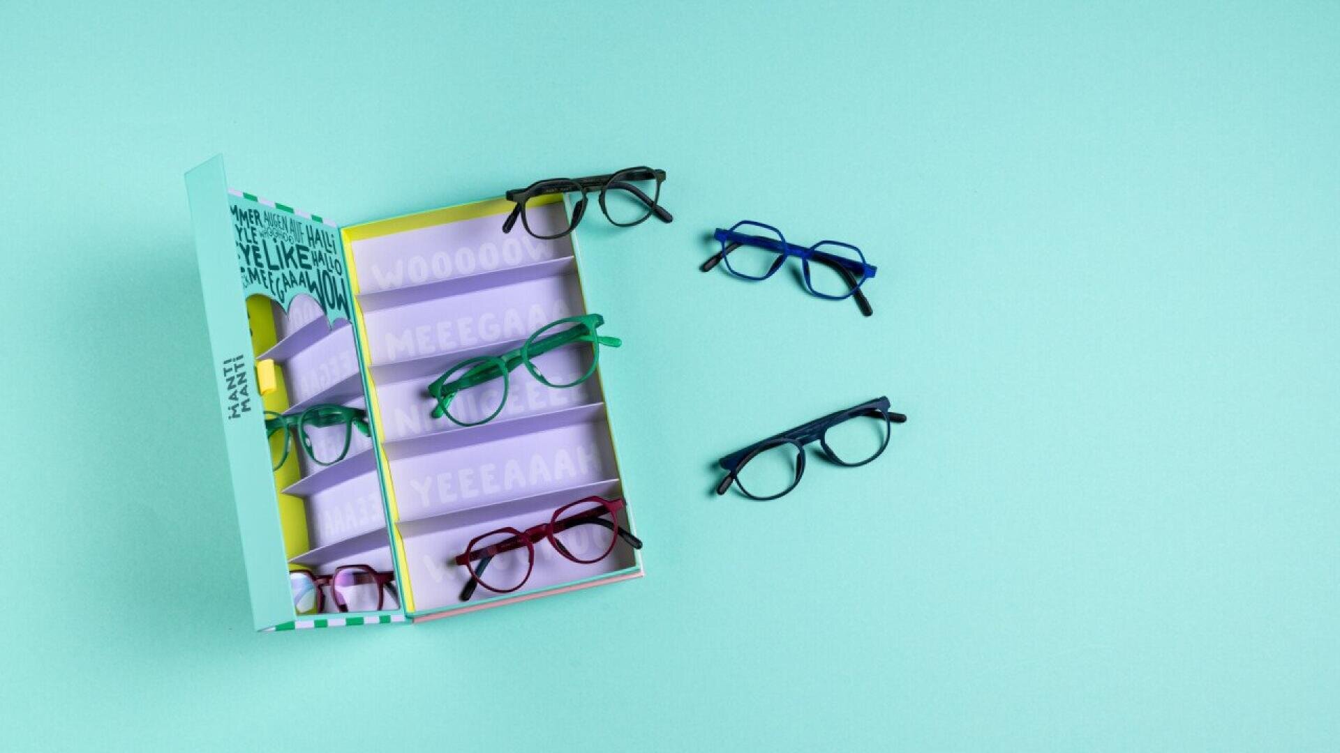 An open box with five pairs of colorful eyeglasses displayed inside on a pastel blue surface, with three more pairs of eyeglasses placed next to the box.