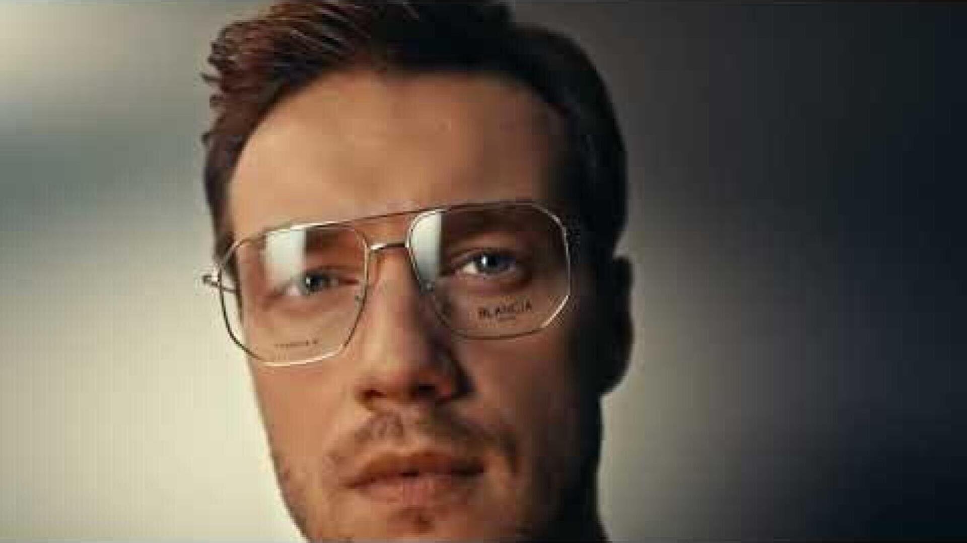 A man with short brown hair and light stubble is wearing large, vintage-style eyeglasses. He is looking directly at the camera with a neutral expression against a blurred background.