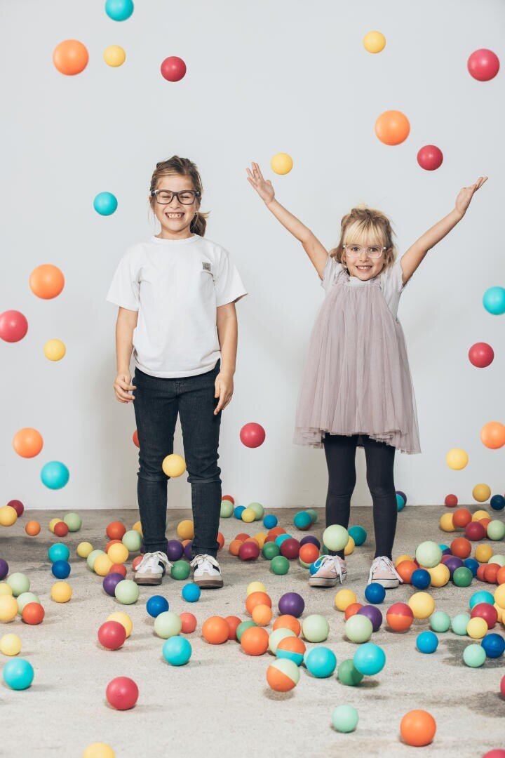 Zwei kleine Kinder stehen umgeben von bunten Plastikbällen. Das Kind links trägt eine Brille und ein weißes Hemd und lächelt mit ausgestreckten Armen. Das Kind rechts trägt ein Kleid und streckt freudig die Arme zwischen den Bällen hervor.