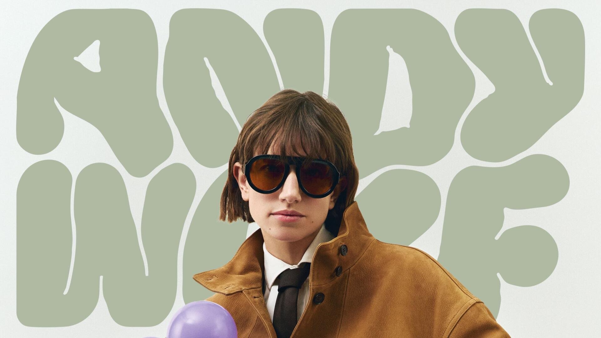 A person wearing large sunglasses, a brown coat and a black tie stands in front of a wall with the words ANDY WAR in bold, bright green, bubbly letters.