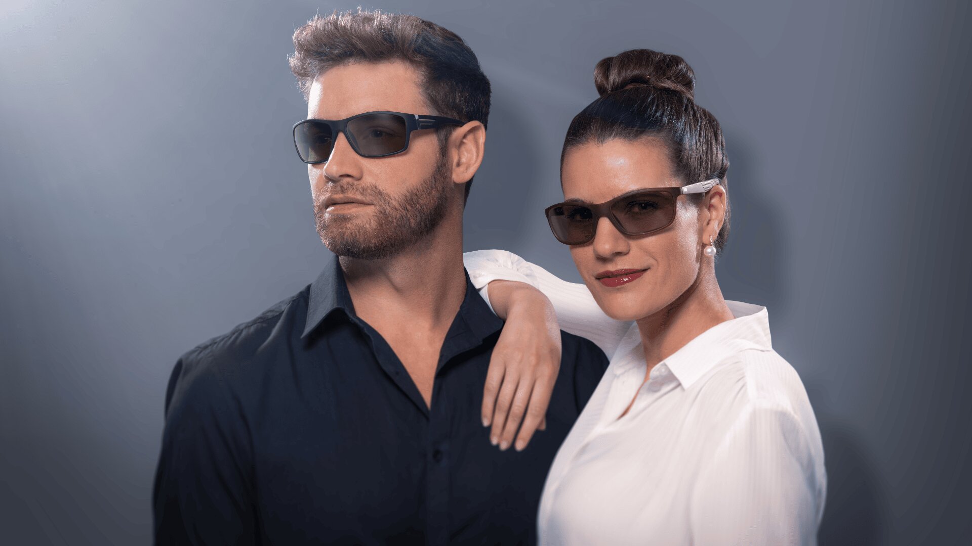 Ein Mann in einem schwarzen Hemd und eine Frau in einer weißen Bluse, beide mit dunkler Sonnenbrille, posieren selbstbewusst vor einem grauen Hintergrund. Die Frau stützt ihre Hand auf die Schulter des Mannes.