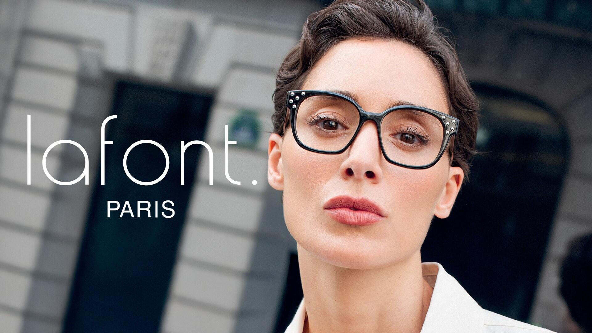 Eine Frau mit kurzen braunen Haaren und einer auffälligen schwarzen Brille blickt selbstbewusst in die Kamera. Das Wort lafont. PARIS wird auf der linken Seite angezeigt, was auf eine Brillenwerbung hindeutet. Im Hintergrund ist ein verschwommenes Stadtgebäude zu sehen.