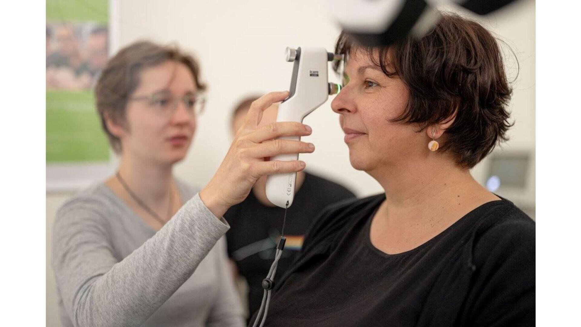 A person uses a handheld device to examine another womans eye in a clinical setting. The woman being examined sits still while the examiner focuses on the procedure.