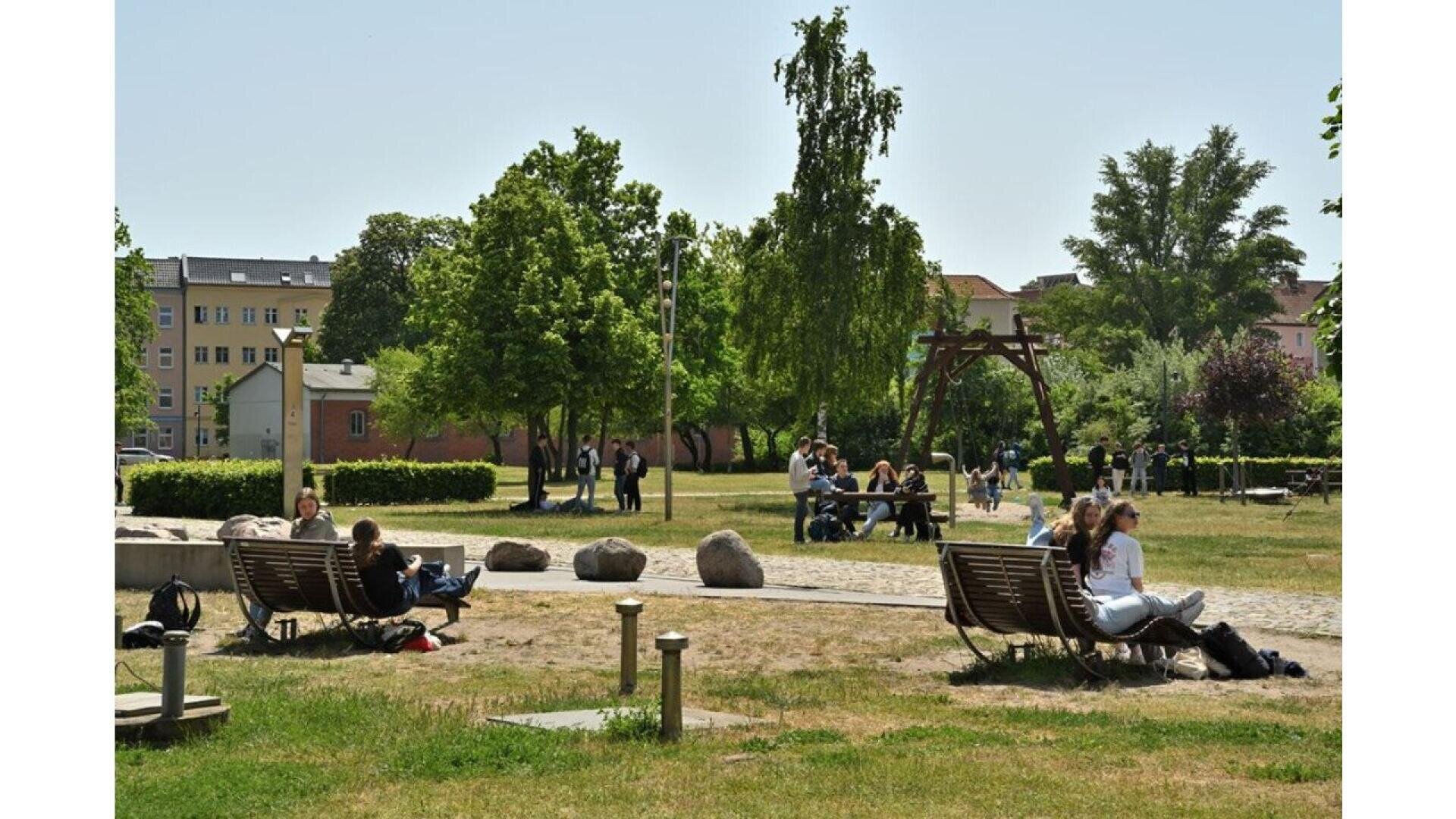 People relax and socialize on benches and grass in a sunny urban park, surrounded by green trees and apartment buildings in the background. Some are talking, reading, or sitting alone.