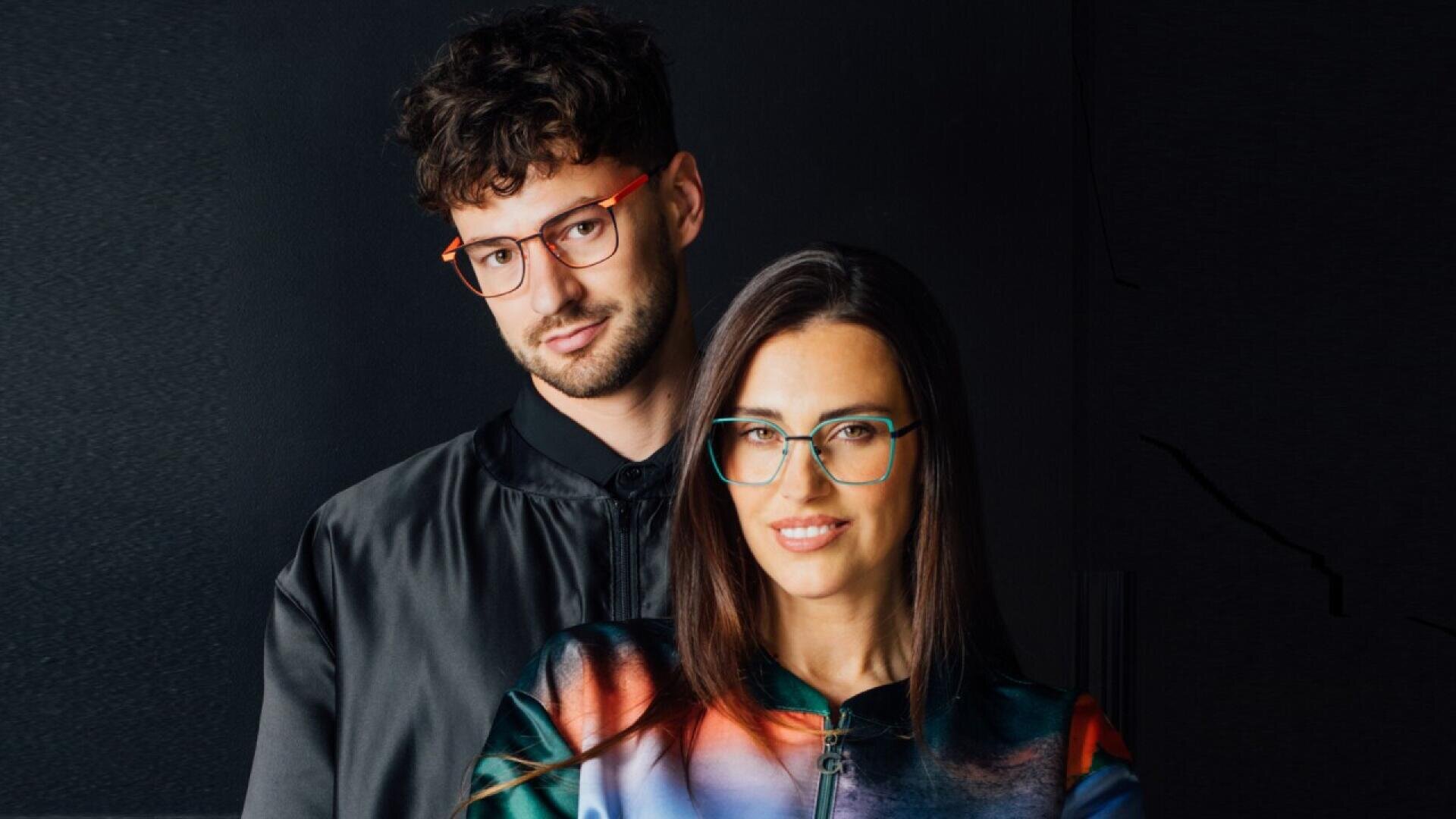A man and a woman wearing glasses stand together against a dark background. The man has curly dark hair and wears a black jacket and orange glasses. The woman has long brown hair, wears colorful clothing, and green glasses.
