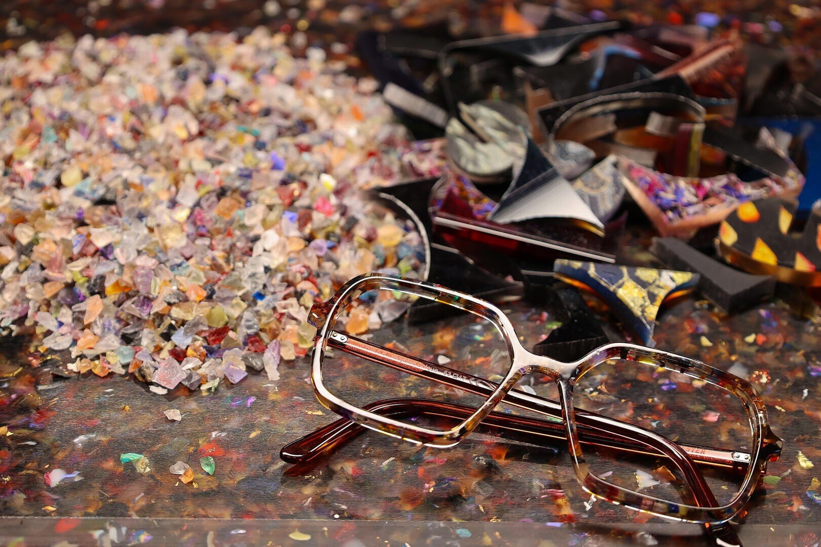 Eine braune, schildpattgemusterte Brille liegt auf einer Glasfläche neben einem Haufen bunter Glasscherben und einem Haufen mehrfarbigen, durchscheinenden Scherbens.