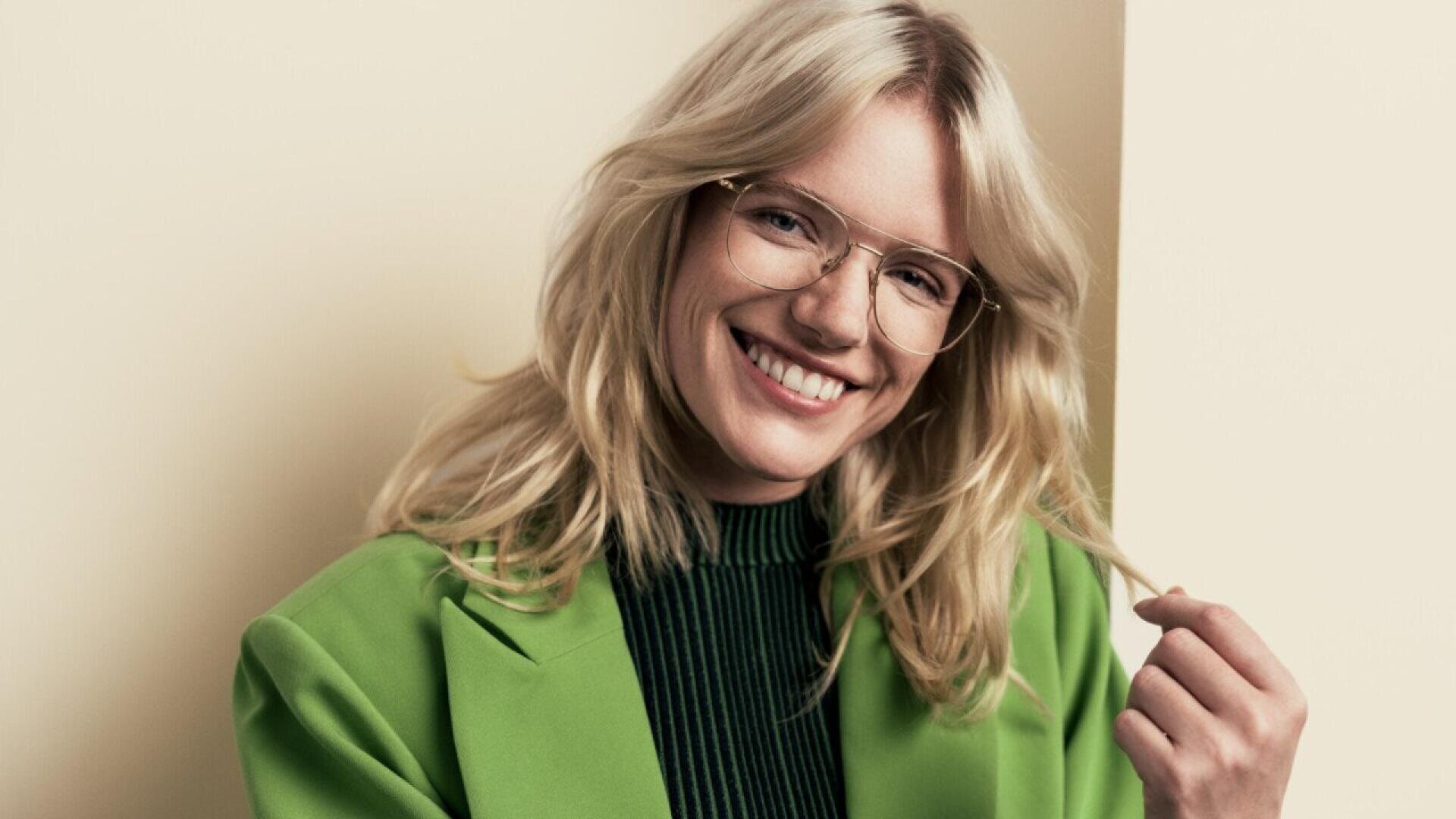 A smiling person with long blonde hair and glasses wears a bright green blazer over a black top, standing against a neutral background and holding a strand of their hair.