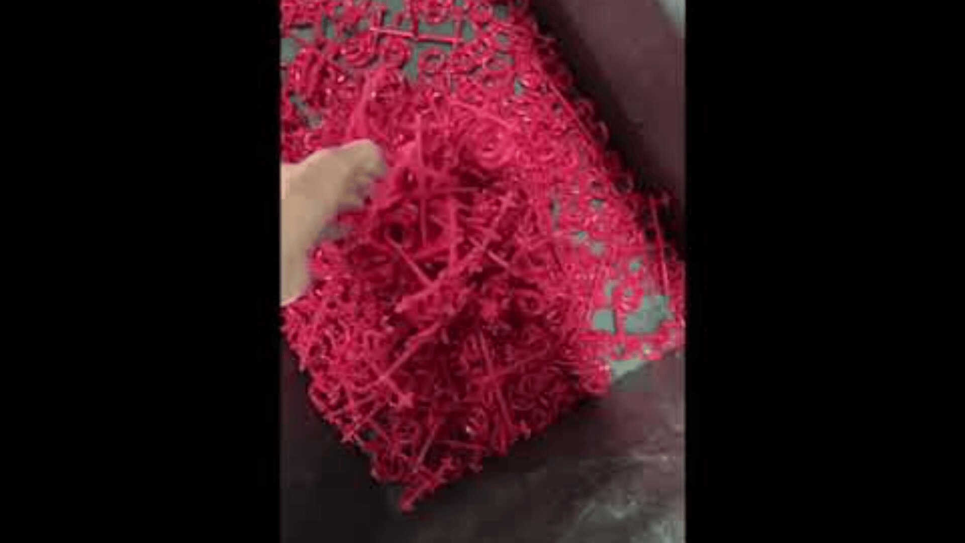 A hand holds a tangled mass of small, red plastic pieces over a container filled with more of the same pieces.
