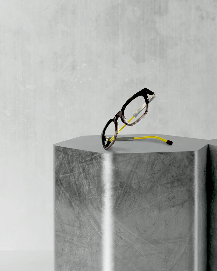Glasses with a black frame and yellow temples resting on a metallic, hexagonal base against a light gray, textured background.