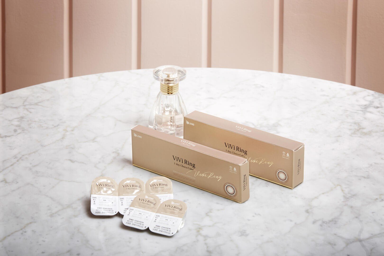 Two beige boxes labeled ViVi Ring stand on a white marble table with several contact lens blisters and a transparent perfume bottle in front of a light pink paneled wall.