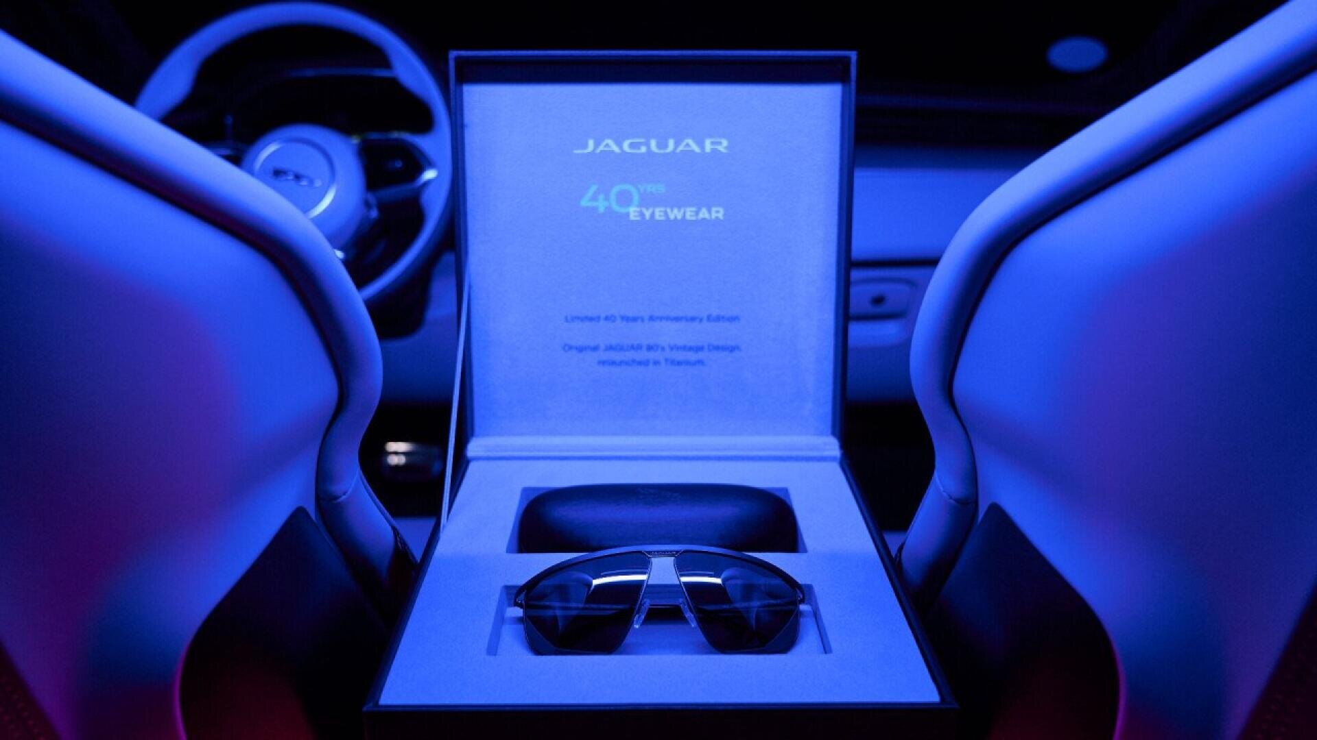 A pair of Jaguar-branded sunglasses sits in an open display box between two illuminated car seats under blue and purple lighting, with a steering wheel visible in the background.