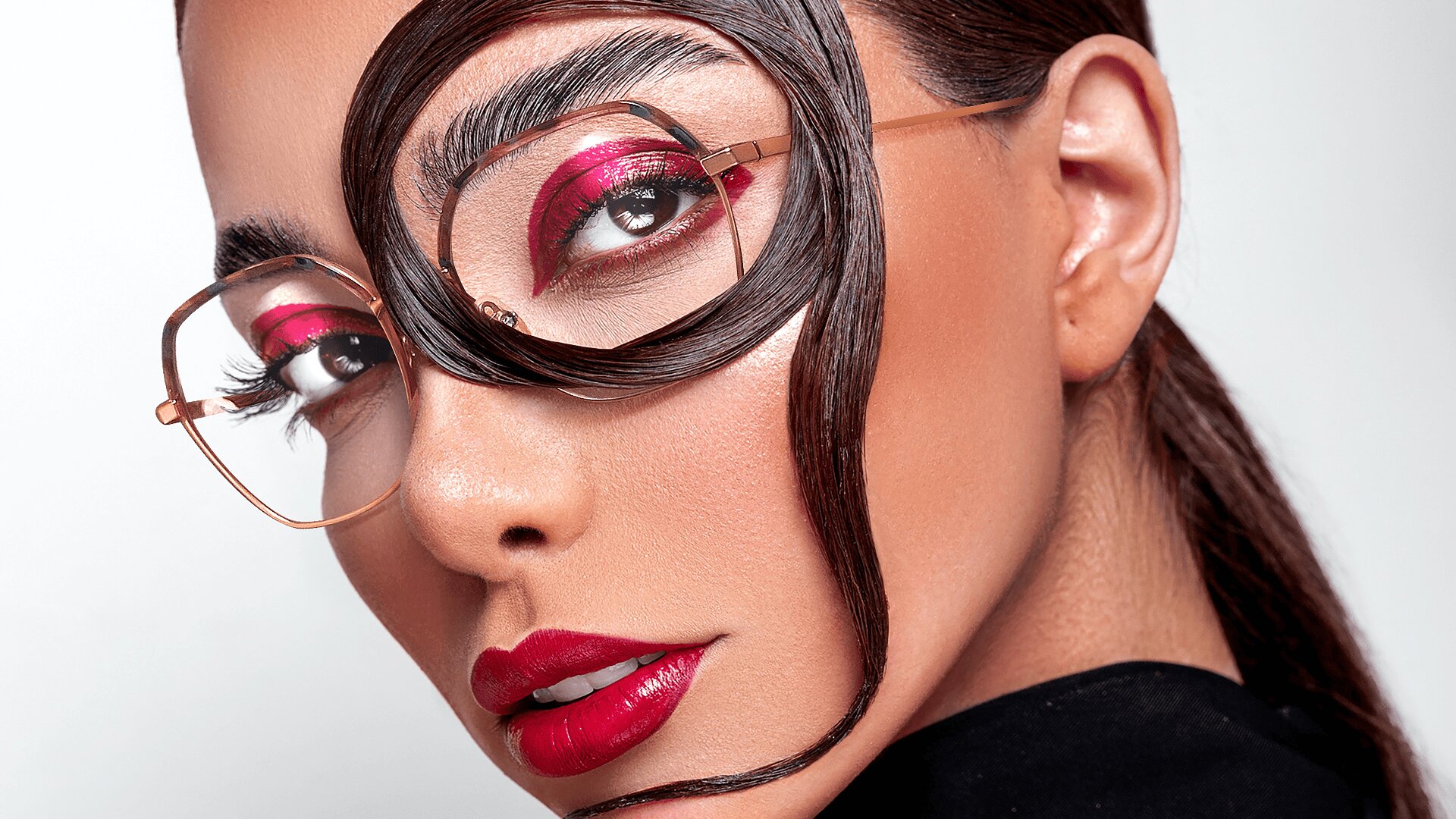 Eine Frau mit kräftigem rosa Lidschatten, glänzendem roten Lippenstift und Brille posiert mit einer kunstvoll über ihr Gesicht und um ihr Auge gelockten Haarsträhne, wodurch ein modischer und kreativer Look entsteht.