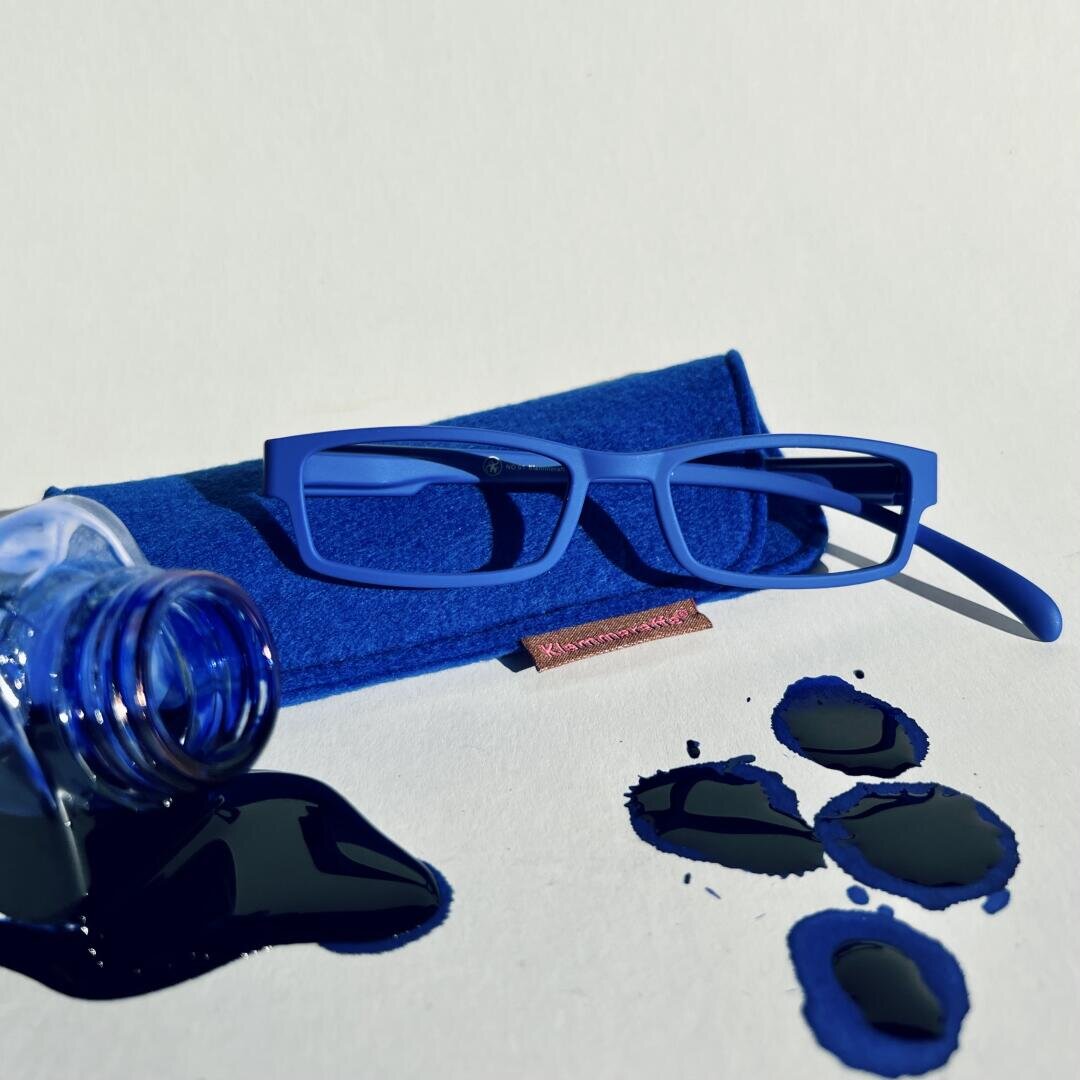 Eine blaue, rechteckige Brille liegt auf einem blauen Etui. Daneben ist eine umgekippte Tintenflasche mit dunkelblauer Tinte verschüttet, die auf einer weißen Oberfläche mehrere Pfützen gebildet hat.