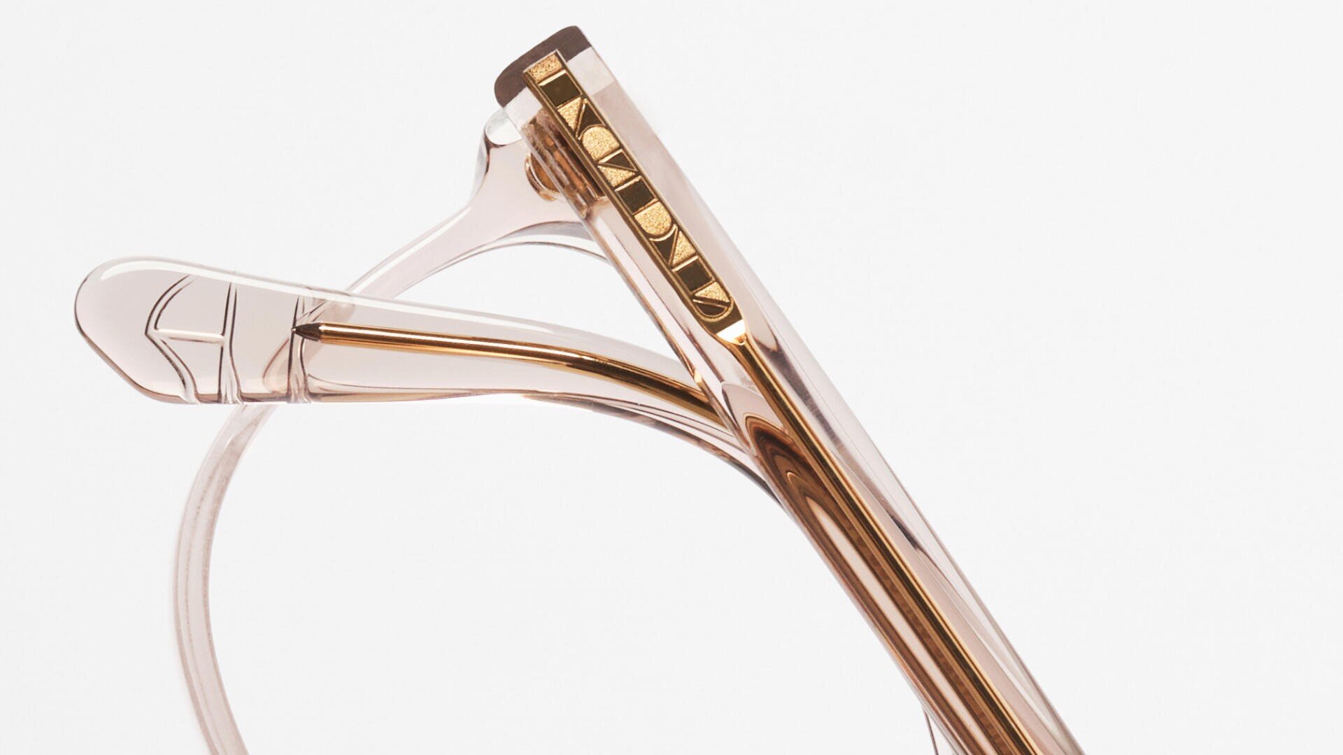 Nahaufnahme des Bügels einer transparenten, hellrosa Brille mit metallisch-goldenem PRADA-Schriftzug und einem geometrischen Designdetail an der Bügelspitze vor einem schlichten weißen Hintergrund.