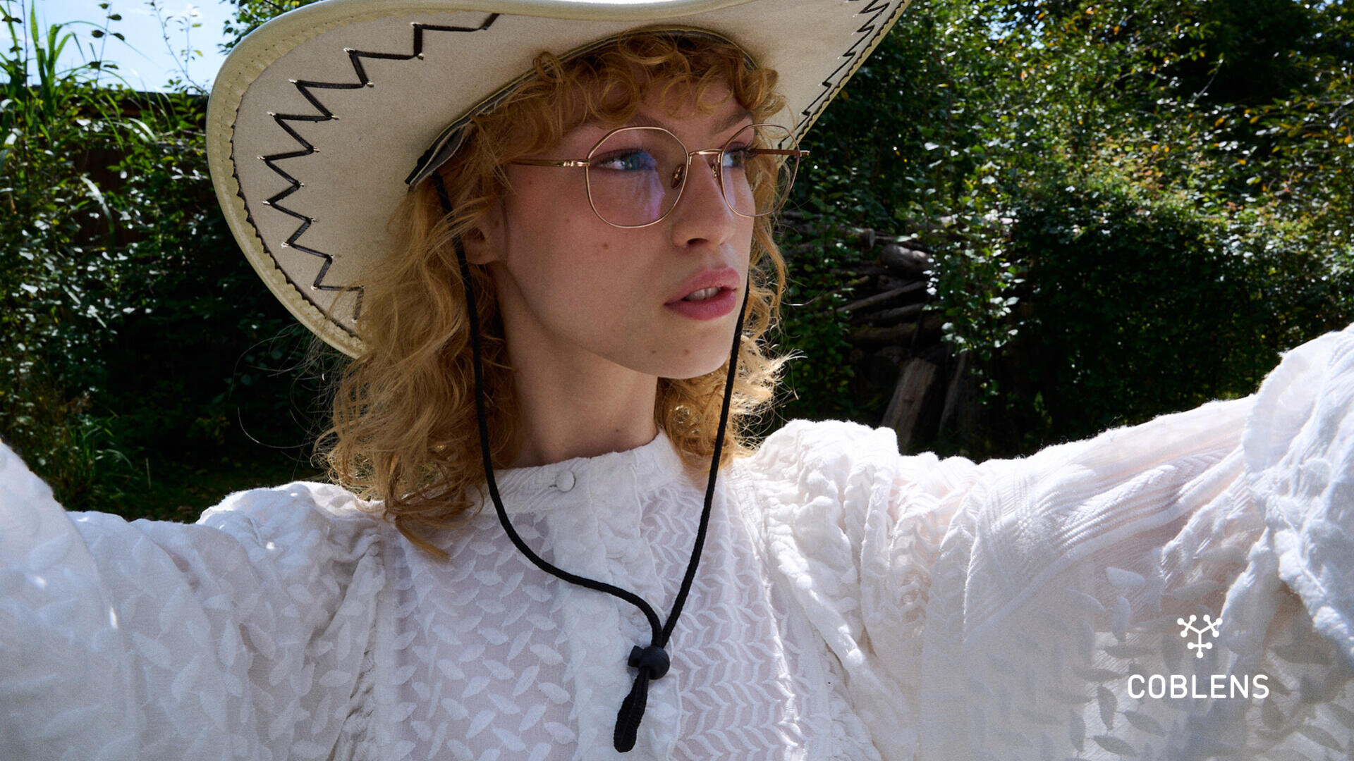 Eine Person mit lockigem blondem Haar, Brille, weißer Bluse und Cowboyhut posiert im Freien in einem Garten. Das Logo von Coblens Eyewear ist unten rechts zu sehen.