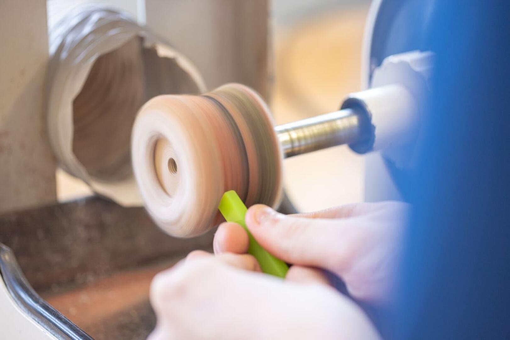 Eine Nahaufnahme von Händen, die ein grünes Objekt gegen eine sich drehende Scheibe einer Polier- oder Schleifmaschine halten, mit Bewegungsunschärfe, die die Scheibe in Aktion zeigt.