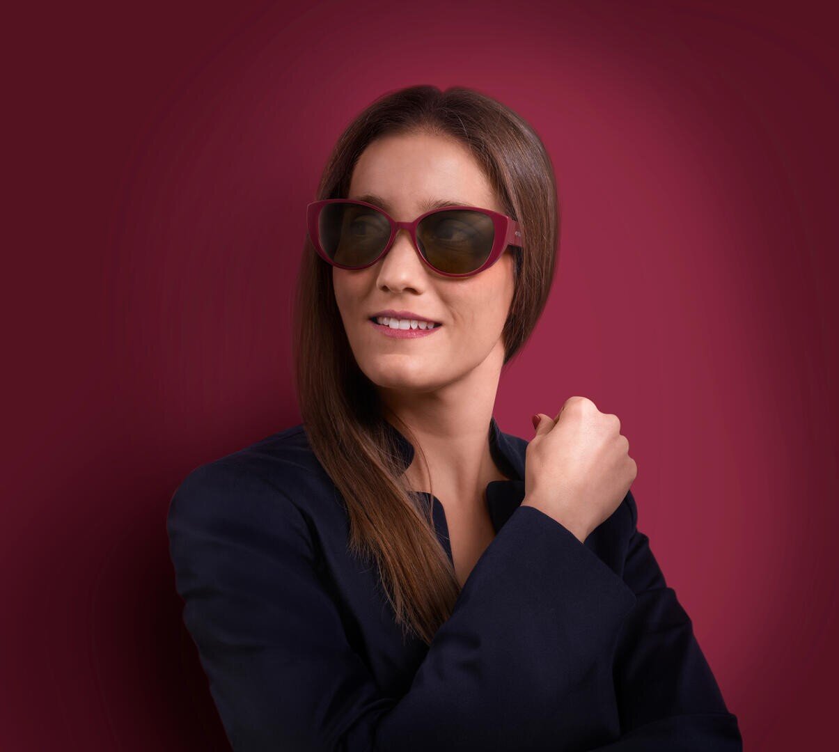 Eine Frau mit langen braunen Haaren, die eine dunkle Sonnenbrille und einen marineblauen Blazer trägt, posiert vor einem einfarbigen kastanienbraunen Hintergrund, lächelt leicht und blickt zur Seite.