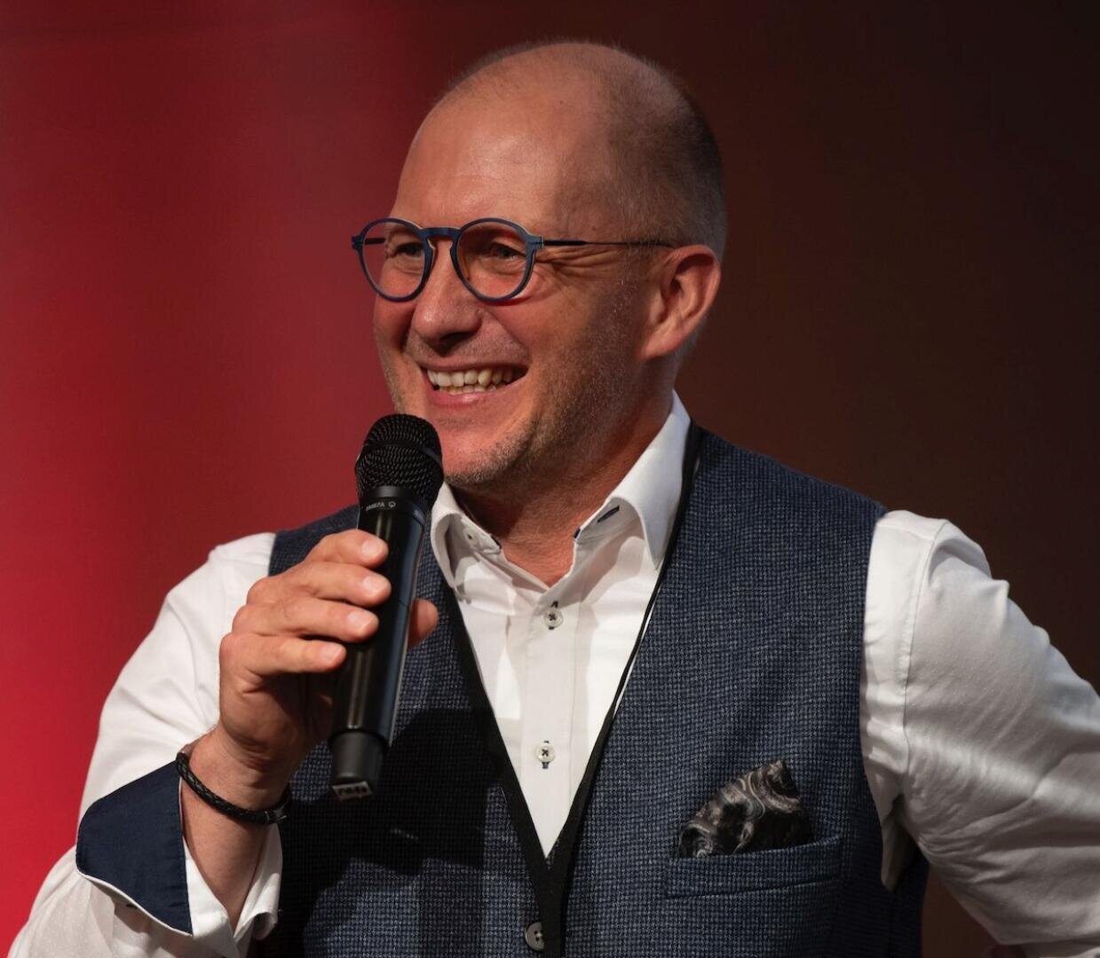 Ein lächelnder, kahlköpfiger Mann mit Brille, weißem Hemd und blauer Weste hält ein Mikrofon, während er vor einem roten und dunklen Hintergrund auf der Bühne spricht.