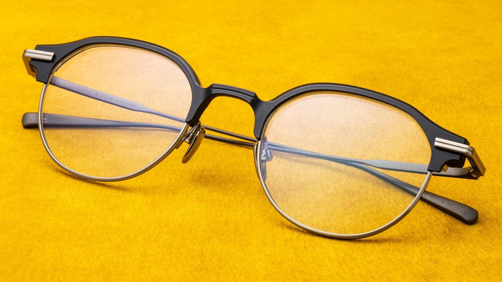 A pair of round, black and silver eyeglasses with clear lenses is placed on a bright yellow surface.