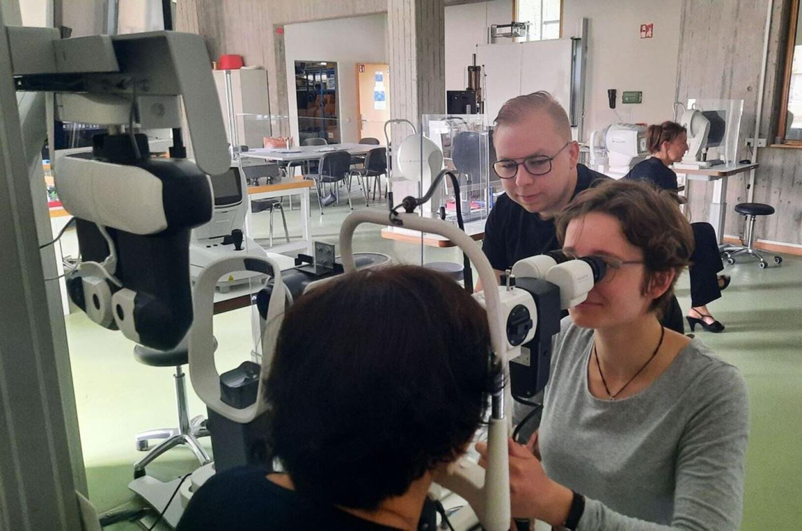 Eine Frau sitzt an einem Augenuntersuchungsgerät, während eine andere Person das Gerät bedient. Eine dritte Person beobachtet sie genau. Der Schauplatz scheint eine moderne Klinik oder ein optometrisches Klassenzimmer mit Geräten und Tischen im Hintergrund zu sein.
