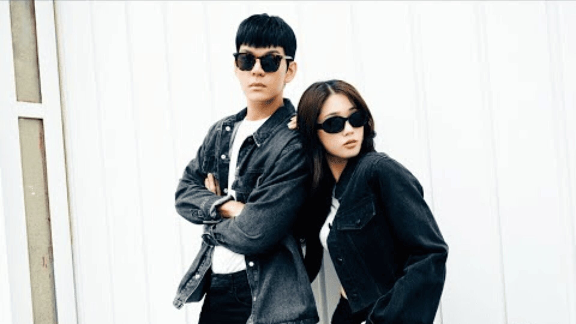 Two people wearing black sunglasses and matching black denim jackets pose confidently together in front of a white wall. One stands with arms crossed, while the other leans in, both looking ahead.
