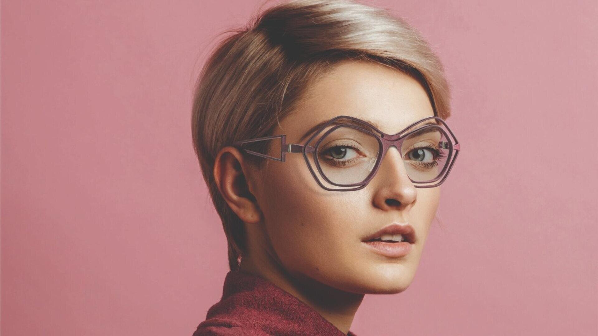 A person with short, light blond hair and large, geometric eyeglasses looks over their shoulder against a pink background, wearing a burgundy top.
