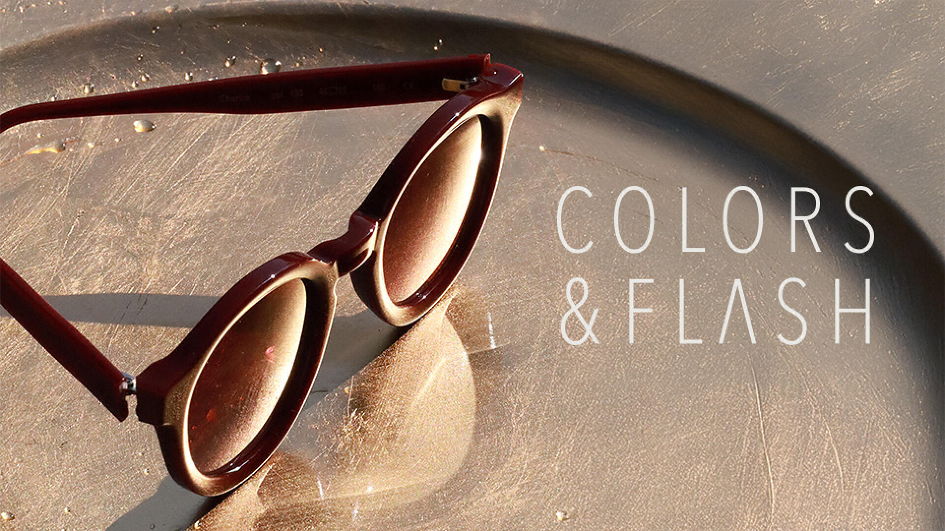 Dark red sunglasses sit on a metallic surface that reflects the light, with the inscription COLORS & FLASH in white lettering on the right-hand side of the picture.