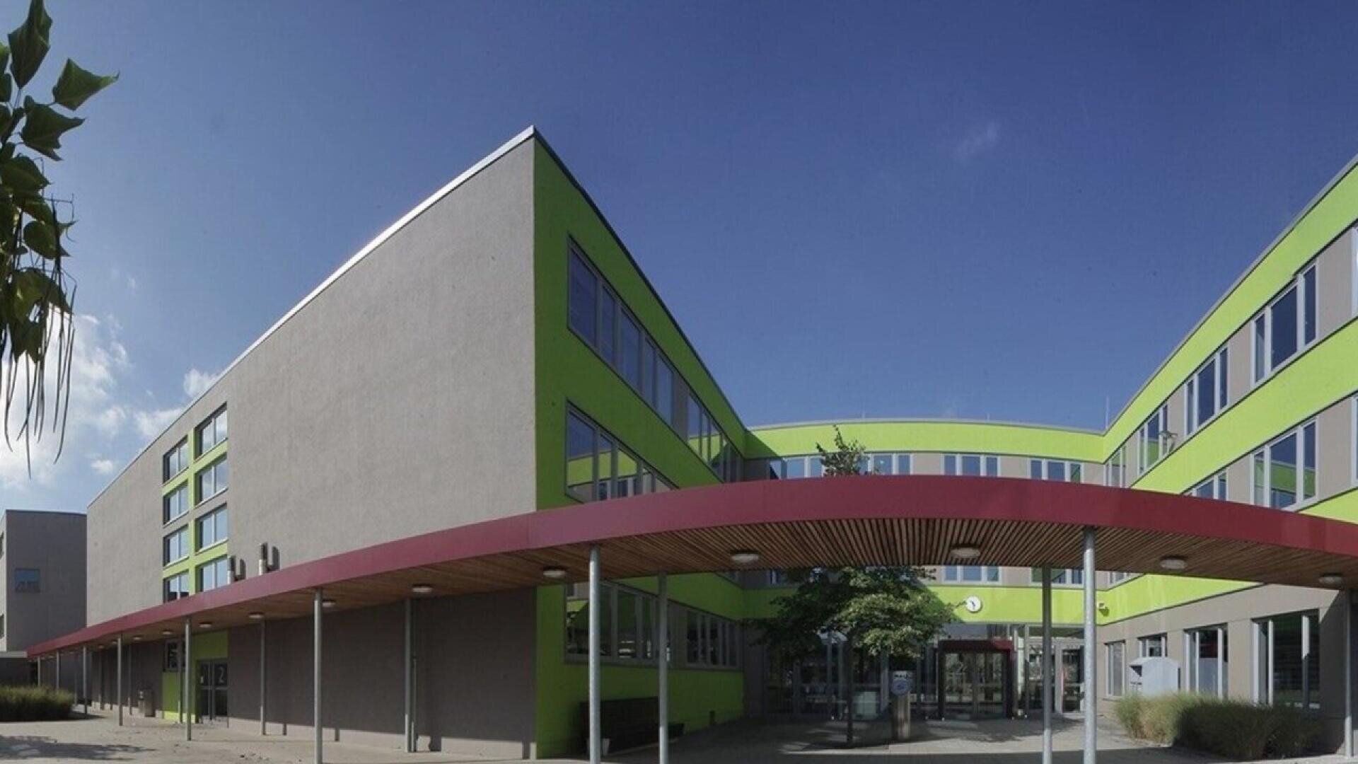 A modern, three-story building with gray and bright green walls, large windows, and a red overhang at the entrance, set against a clear blue sky.