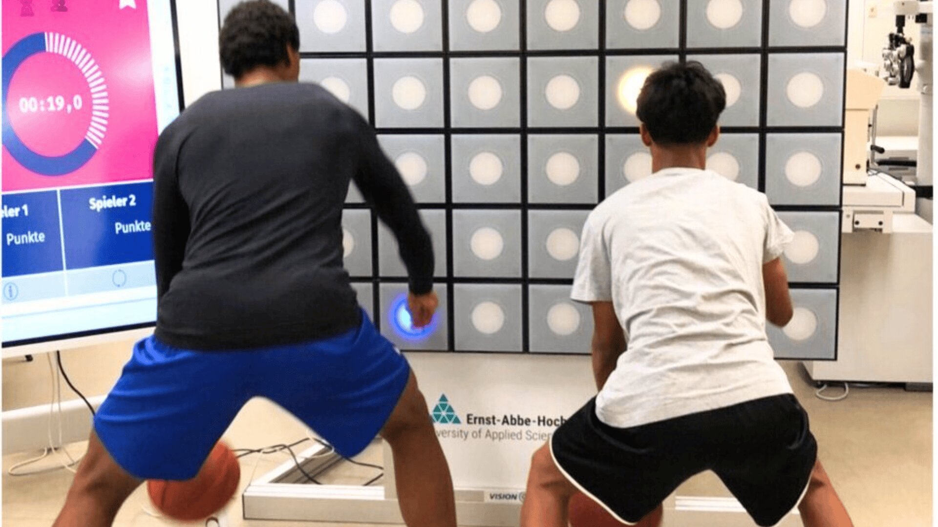 Zwei Personen in Sportkleidung dribbeln Basketbälle, während sie vor einer großen, an der Wand montierten Reaktionstrainingsplatte mit beleuchteten Kreisen stehen und versuchen, die beleuchteten Punkte während einer Übung zu berühren.