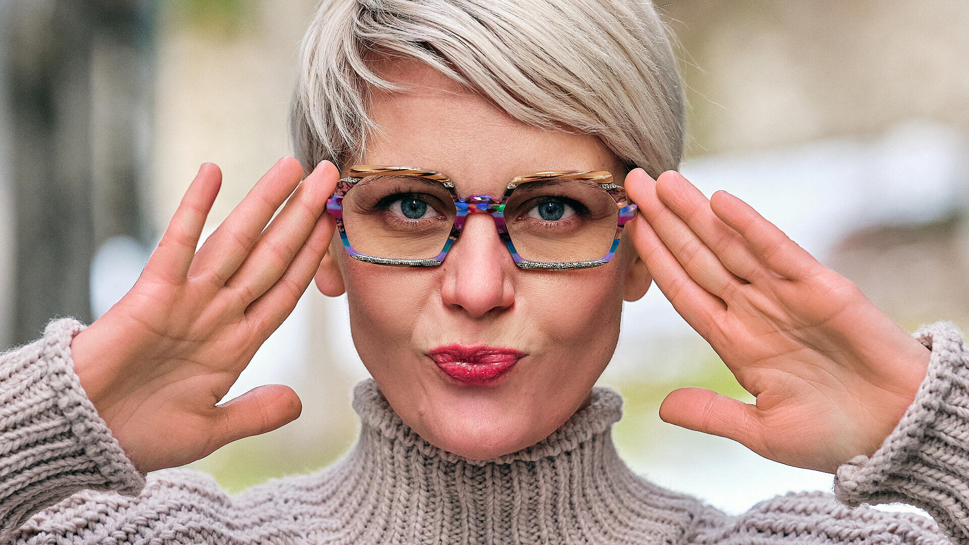 Eine Person mit kurzen blonden Haaren und bunter Brille posiert verspielt, hält die Hände vors Gesicht und schürzt die Lippen. Sie trägt einen dicken, hellen Strickpullover.