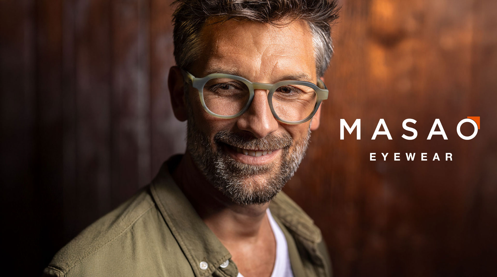 Ein lächelnder Mann mit grauem Haar und Bart trägt eine hellgrüne Brille und ein hellbraunes Hemd und steht vor einem dunklen Holzhintergrund. Neben ihm erscheint der Text MASAO EYEWEAR.