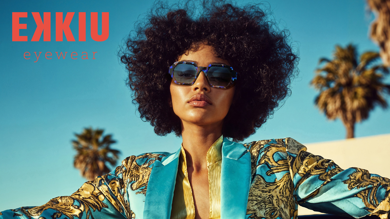 Eine stilvolle Frau mit lockigem Haar tr&auml;gt eine gro&szlig;e Sonnenbrille und eine gewagte, bunte Jacke im Freien, mit Palmen und blauem Himmel im Hintergrund. Der Text EKKIU eyewear erscheint in der oberen linken Ecke.