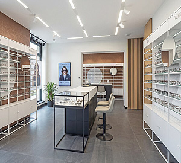 Modern eyewear store with white shelves displaying many pairs of glasses, a central counter with stools, a wall mirror, bright lighting and a large poster of a woman wearing glasses.