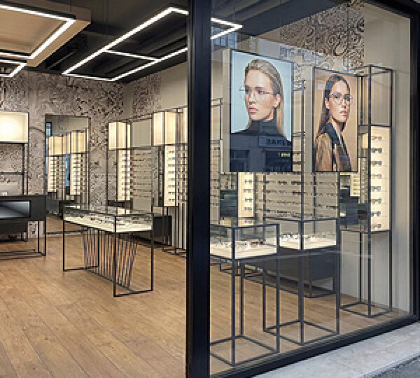 A modern glasses store with glass showcases full of glasses and two large posters of women wearing fashionable glasses on the wall, viewed from outside through a large window.